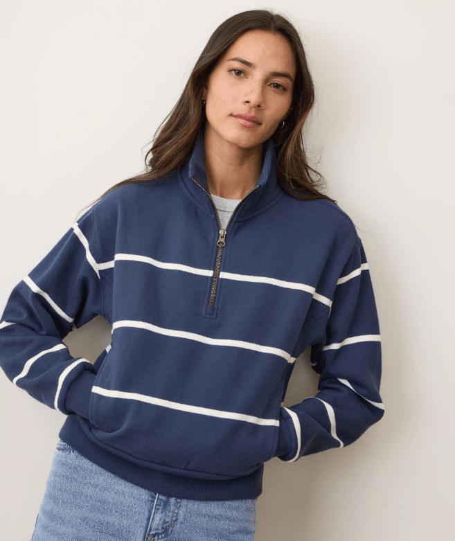 A woman wearing a navy half zip pullover with white stripes and blue jeans on a white background