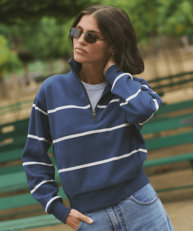 A woman outside at a park wearing a navy half zip pullover with white stripes, blue jeans, and sunglasses