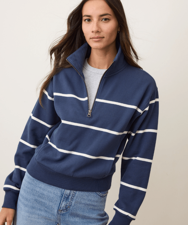 A woman wearing a navy half zip pullover with white stripes and blue jeans on a white background