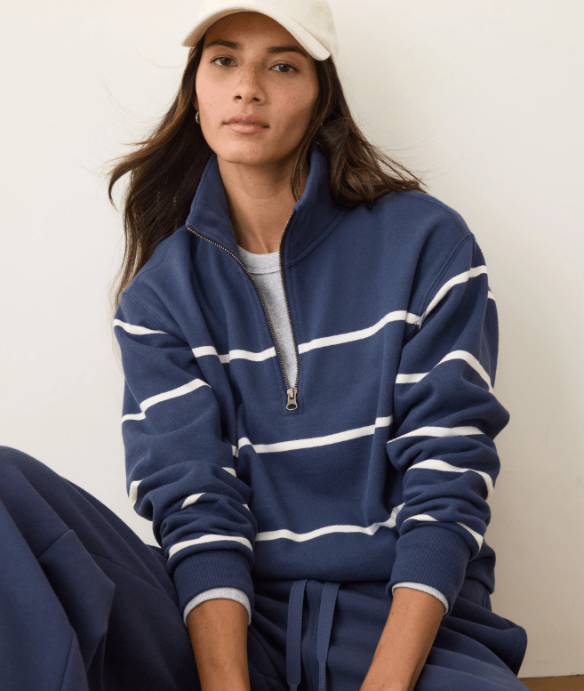 A woman wearing a navy half zip pullover with white stripes, matching navy sweatpants, and a hat on a white background