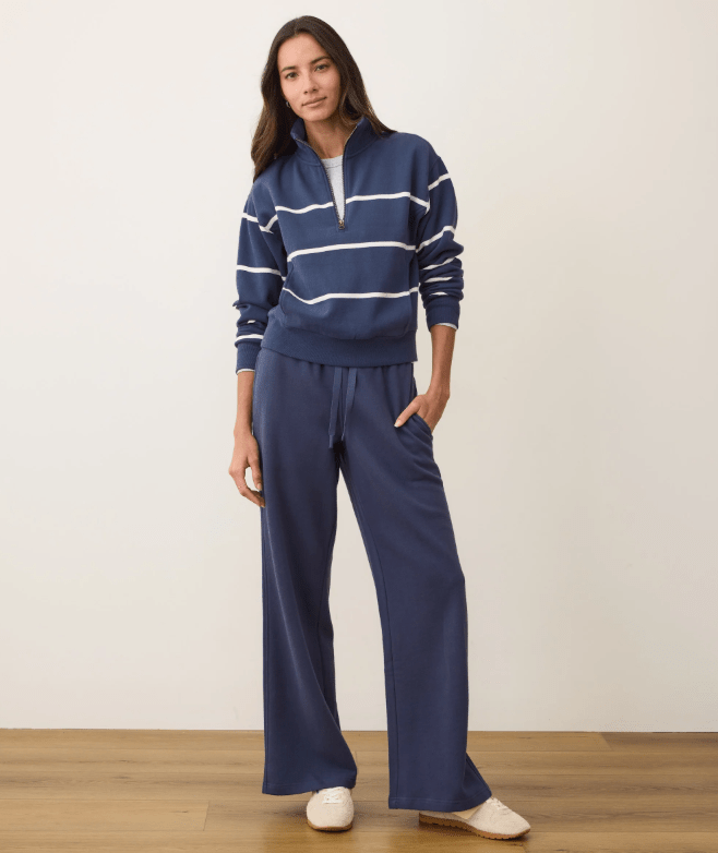 A woman wearing a navy half zip pullover with white stripes and matching navy sweatpants on a white background