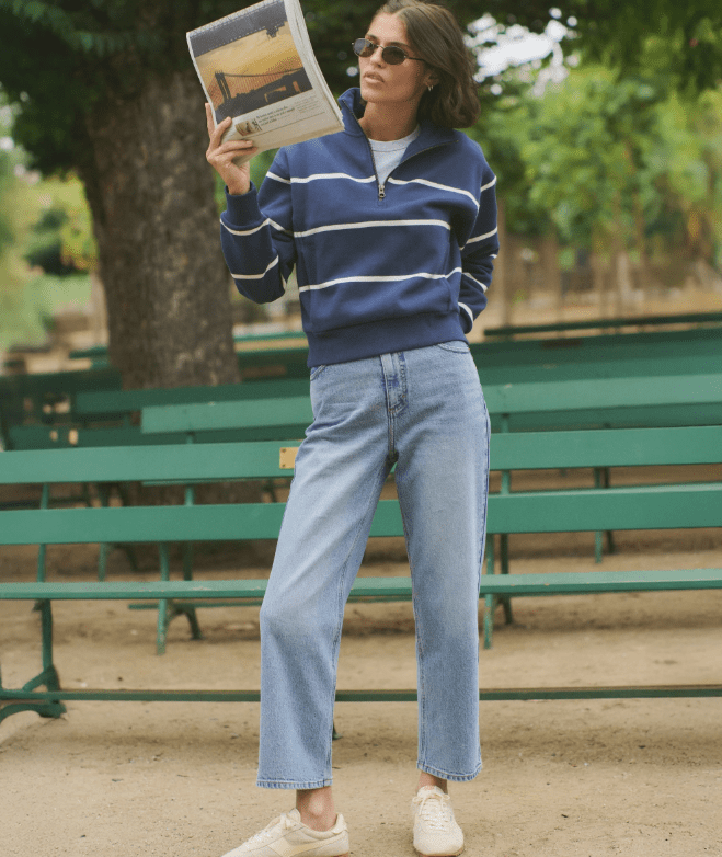 A woman outside at a park wearing a navy half zip pullover with white stripes and blue jeans