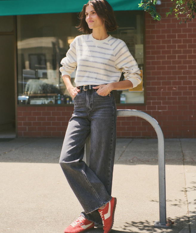 Woman wearing a black and white striped sweater, jeans, and red shoes standing on a sidewalk.