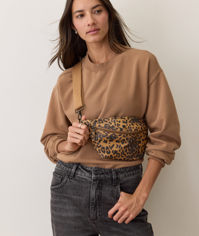 Woman wearing a brown sweatshirt and holding a leopard print fanny pack against a plain background