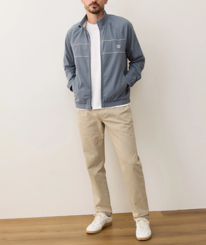 Person wearing a blue windbreaker, white shirt, beige pants, and white sneakers against a plain background