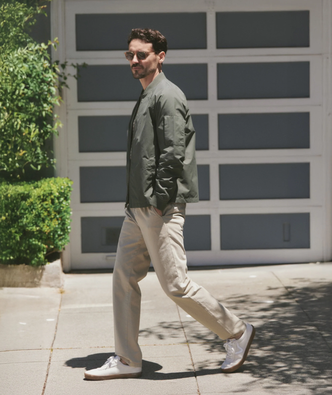 Man walking outdoors in front of a garage door wearing a dark green bomber jacket and beige pants with white shoes