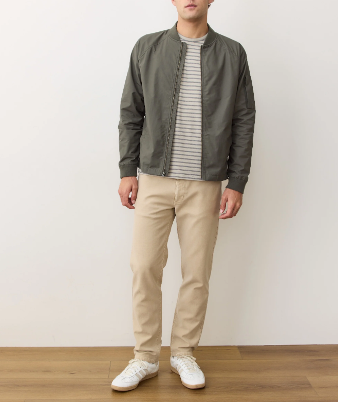 Person wearing a green bomber jacket, striped shirt, beige pants, and white sneakers on a plain background