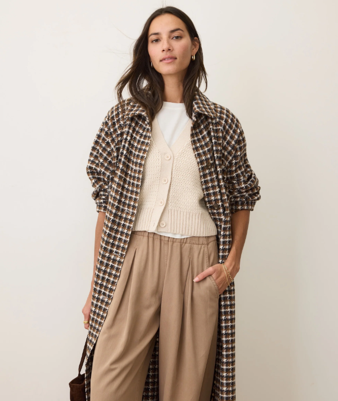 Woman wearing a brown houndstooth coat over a white sweater and beige pants on a plain background