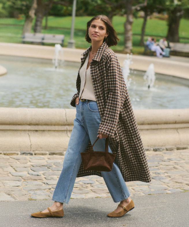 Woman walking in a park with a fountain, wearing a brown houndstooth coat and jeans.