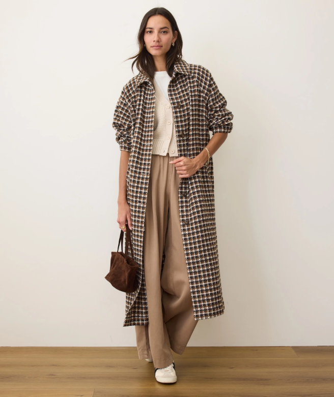Woman wearing a long brown houndstooth coat over a beige dress, holding a brown handbag against a white background.