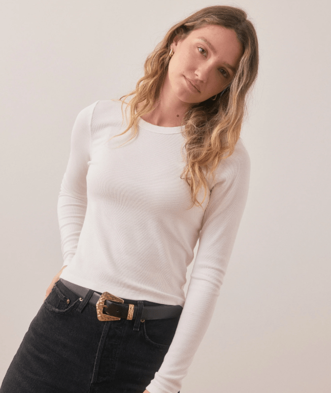 Woman wearing a white long-sleeve top and black jeans against a plain background