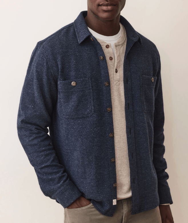 Man wearing a blue herringbone overshirt over a beige sweater on a plain background