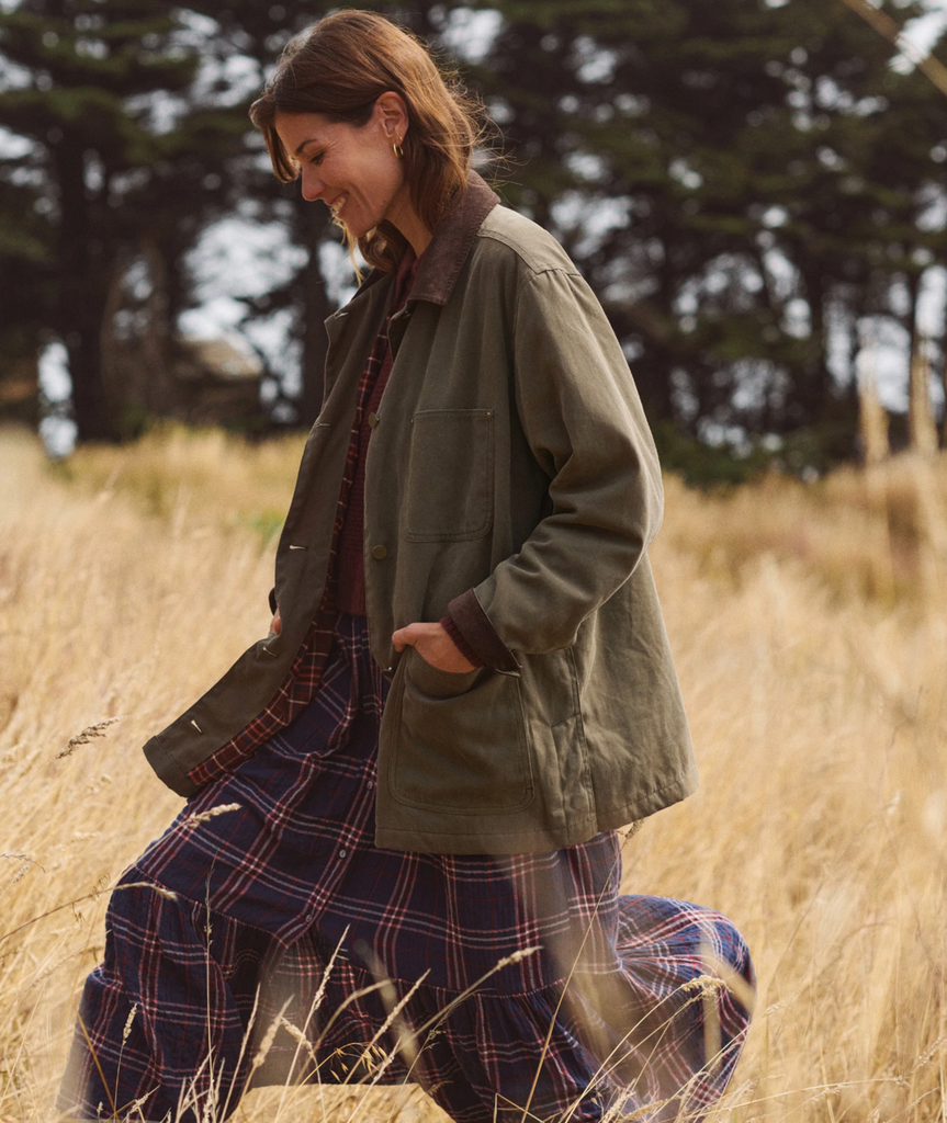 Woman in a green jacket and a plaid dress walking through a field with trees in the background