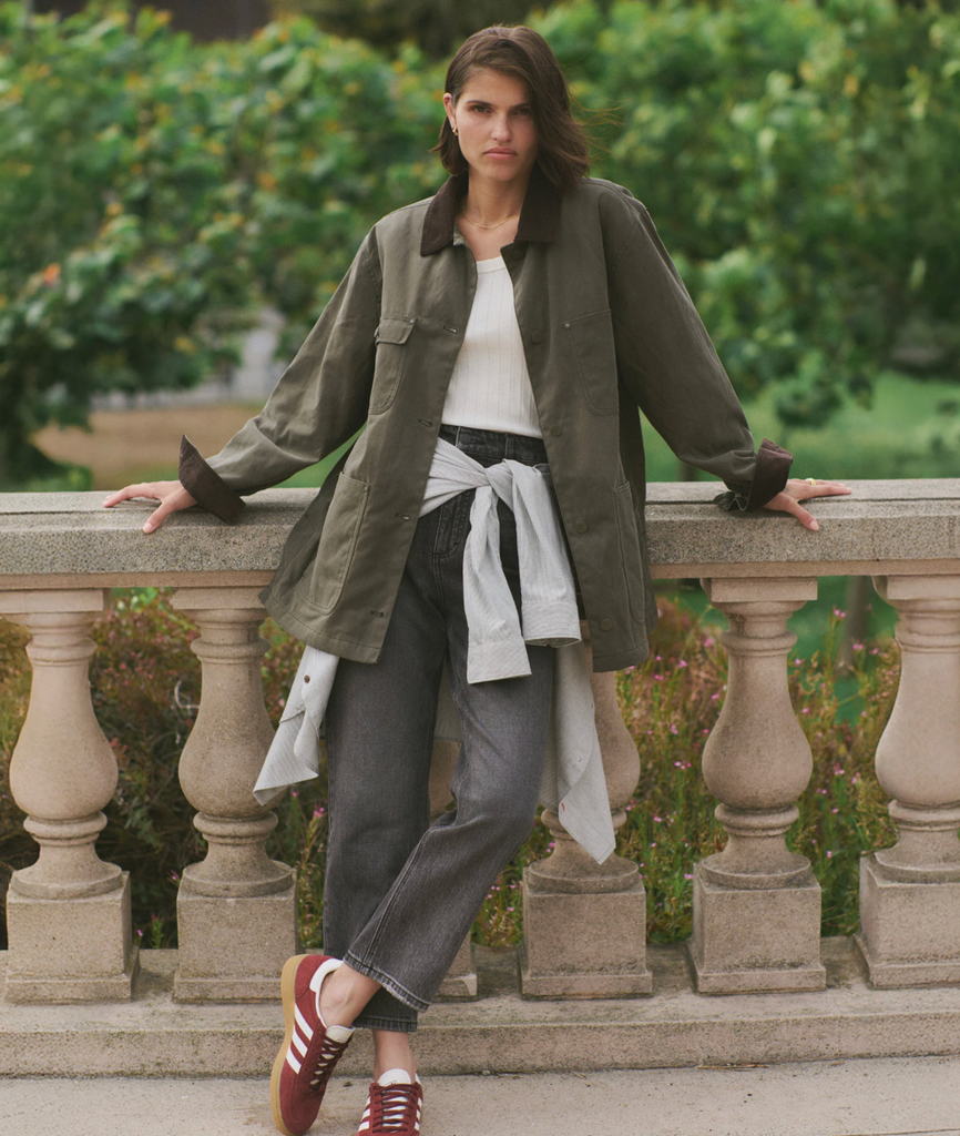 Woman wearing a green jacket, standing on a stone ledge with greenery in the background