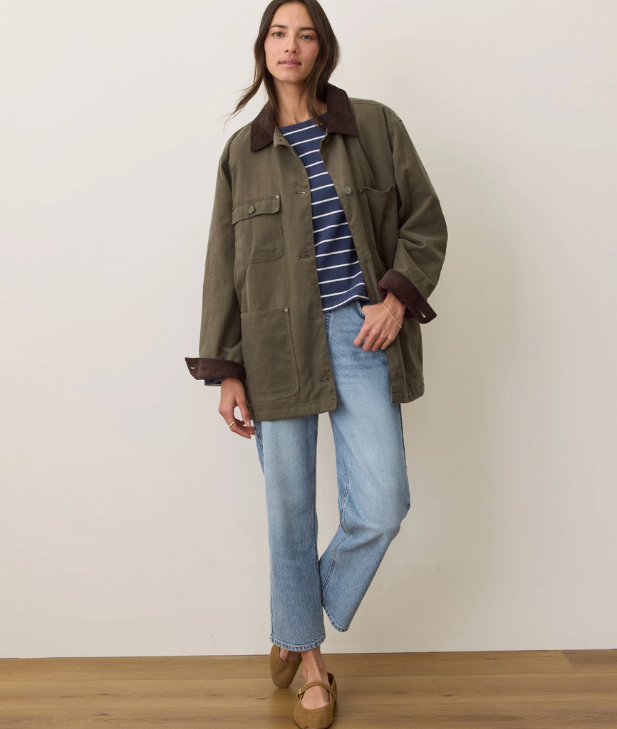 Woman wearing an olive green jacket, striped shirt, and blue jeans against a plain background