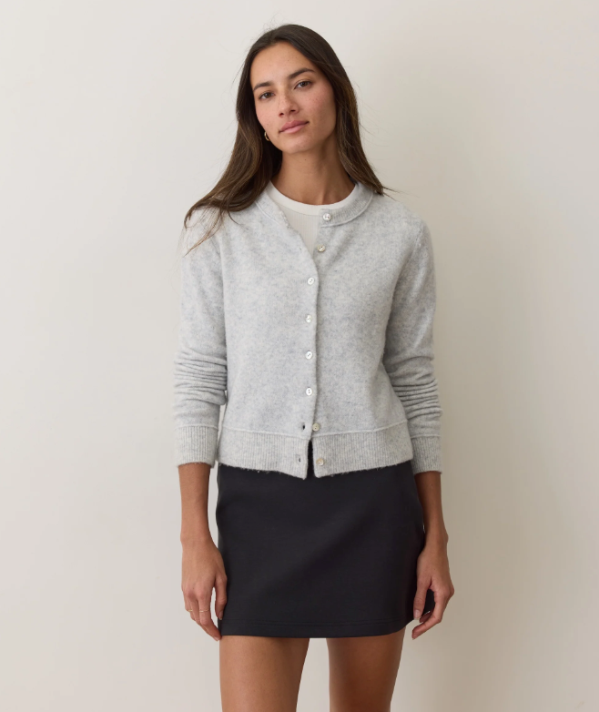 Woman wearing a gray cardigan over a white shirt with a black skirt on a plain background