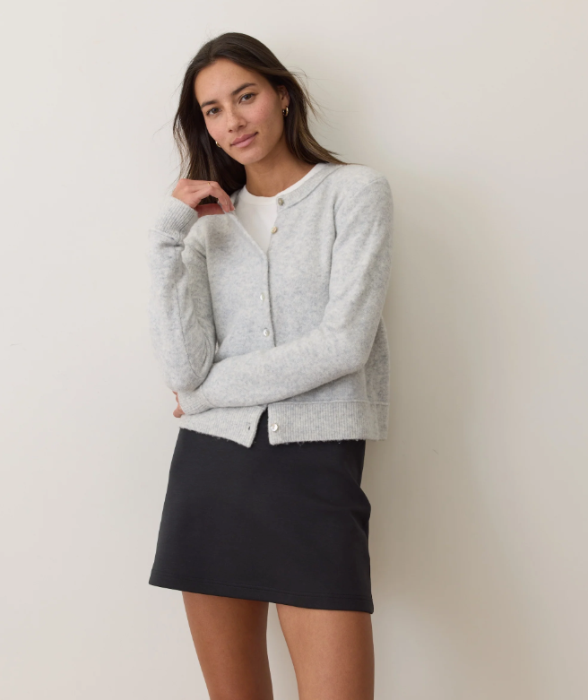 Woman wearing a gray cardigan over a white shirt and black skirt against a plain background