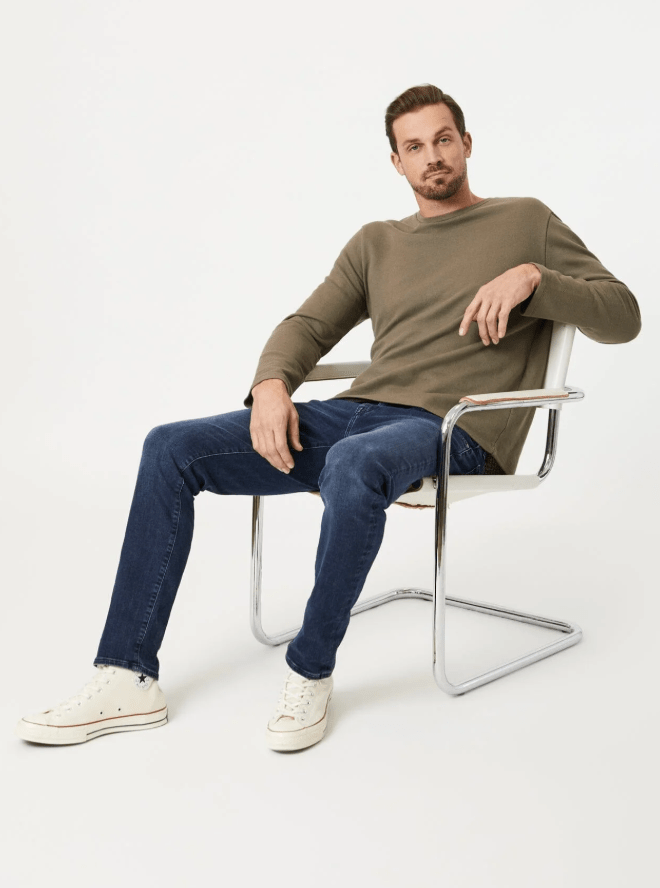 Man sitting on a chair wearing a green sweater and blue jeans against a white background