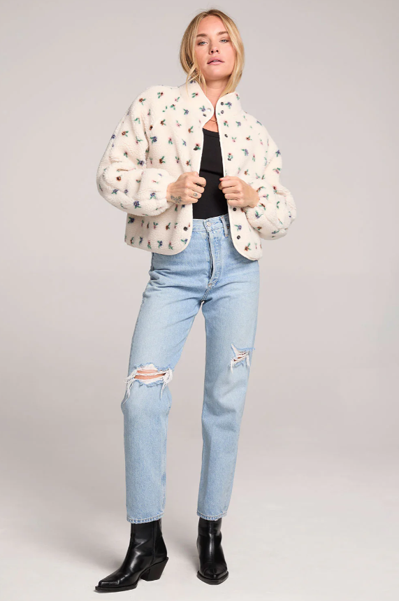 Woman wearing a fluffy floral patterned jacket, black top, light blue jeans, and black boots on a plain background