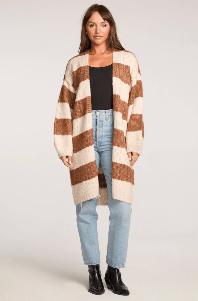 Woman wearing a long brown and cream striped cardigan, light blue jeans, and a black shirt on a white back ground