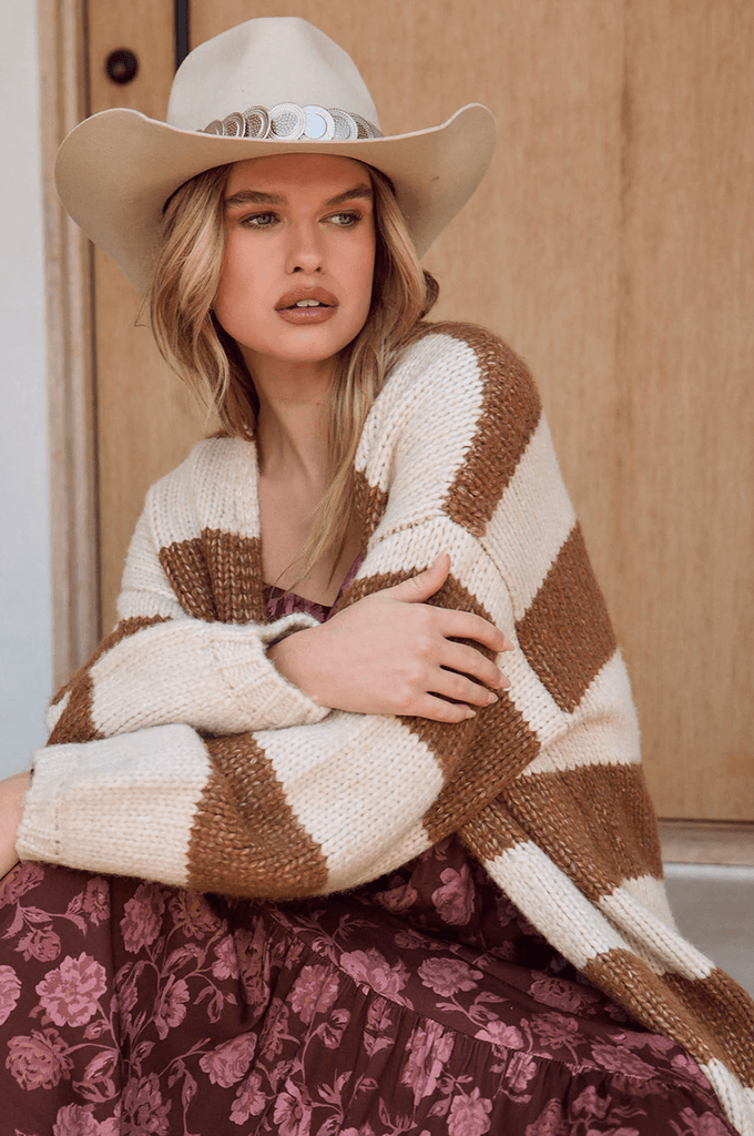 Woman wearing a long brown and cream striped cardigan and a cowboy hat on a porch