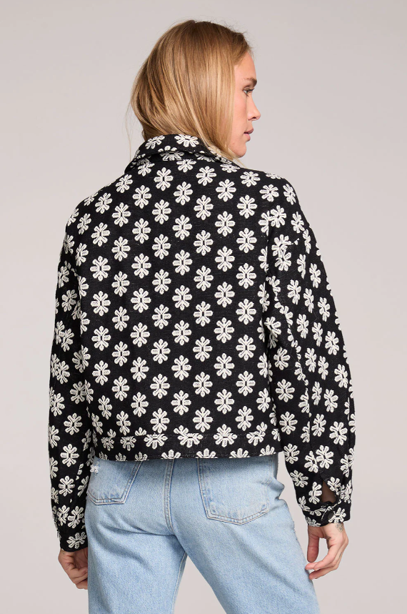 Person wearing a black jacket with white floral pattern and light blue jeans on a plain background