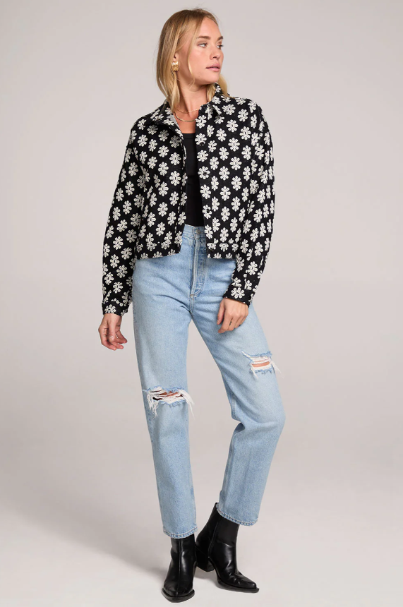 Woman wearing a black and white floral jacket, black top, light blue jeans, and black boots on a plain background