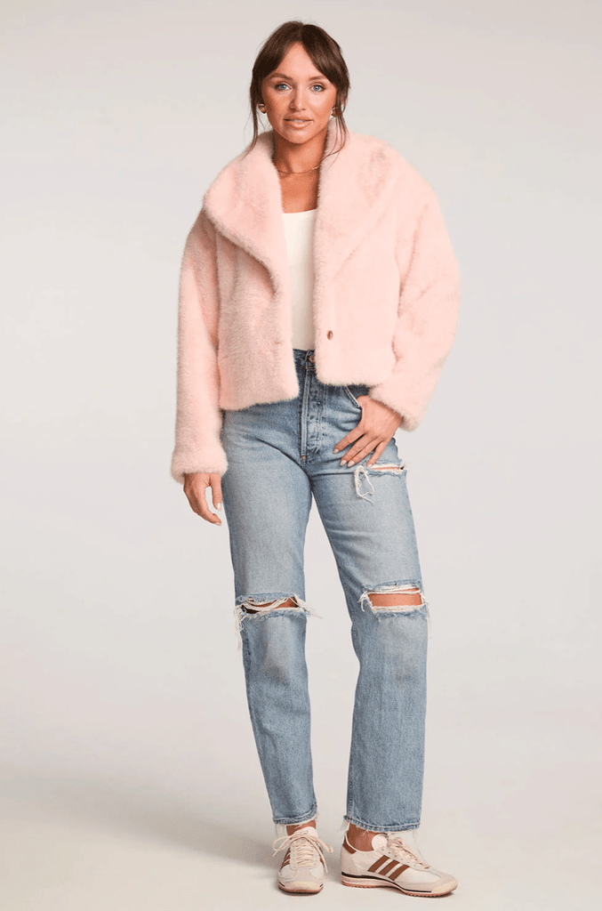 Woman wearing a pink coat, white top, and ripped jeans on a plain background