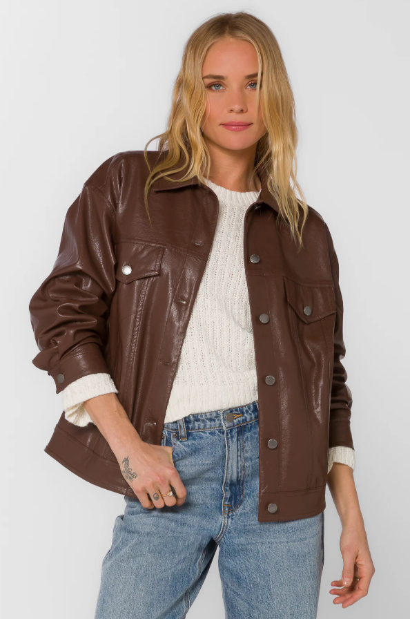 Woman wearing a brown faux leather jacket over a white sweater and blue jeans on a plain background