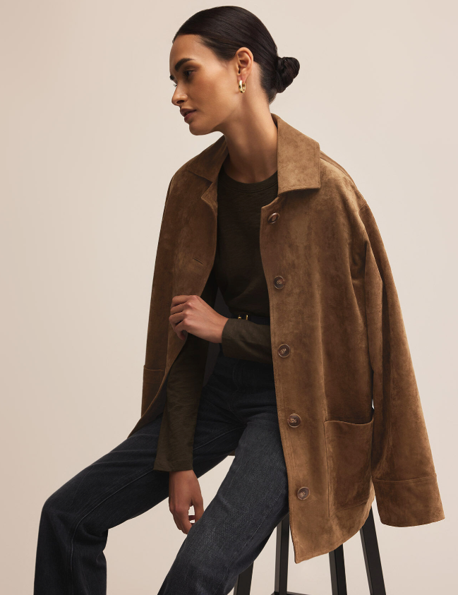 Woman wearing a brown faux suede coat sitting on a stool against a plain background