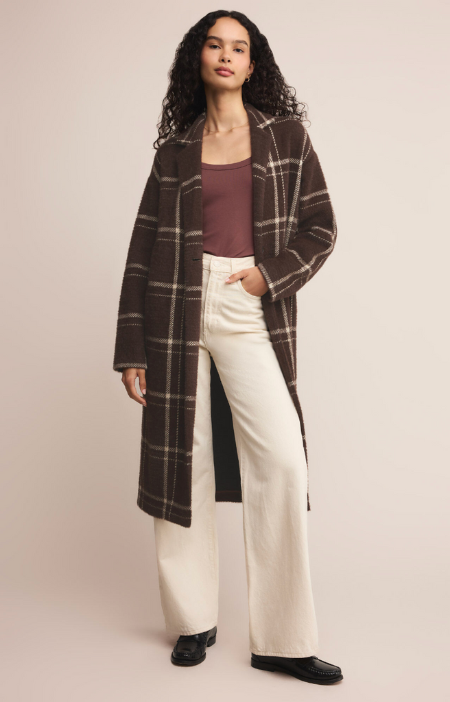 Woman wearing a brown plaid coat, purple top, and white pants on a beige background