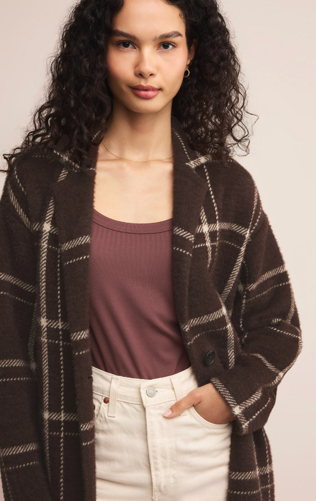 Woman wearing a brown plaid coat over a purple top and white pants on a plain background