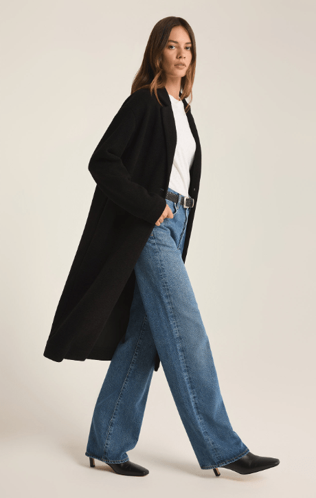 Woman wearing a long black coat, white shirt, and blue jeans on a beige background