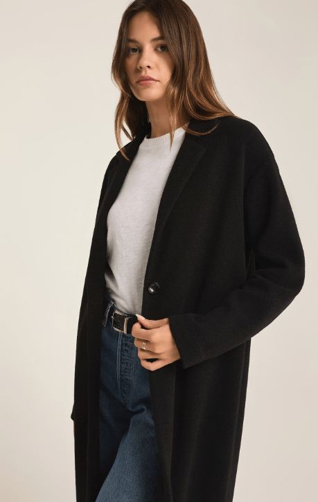 Woman wearing a long black coat over a white shirt and blue jeans on a plain background